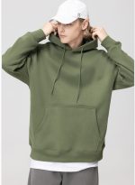 A hooded sweatshirt in olive green, featuring a front pocket and drawstrings. The model wears a white cap.