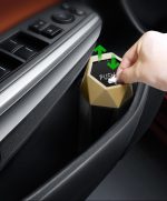 A hexagonal, metallic car trash can with a push lid, featuring black and gold colours, being interacted with by a hand.