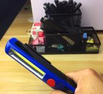 A handheld work light in blue with a yellow LED strip, positioned in front of a pen holder filled with various items.