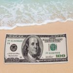 A beach towel featuring a large printed image of a hundred-dollar bill, set on a sandy beach with water.