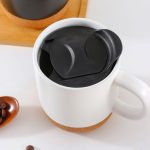 A ceramic mug with a cork base, featuring a black lid, filled with coffee beans on a wooden surface.