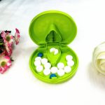 A green plastic pill cutter and pill box, containing white and blue pills, with floral decorations nearby.