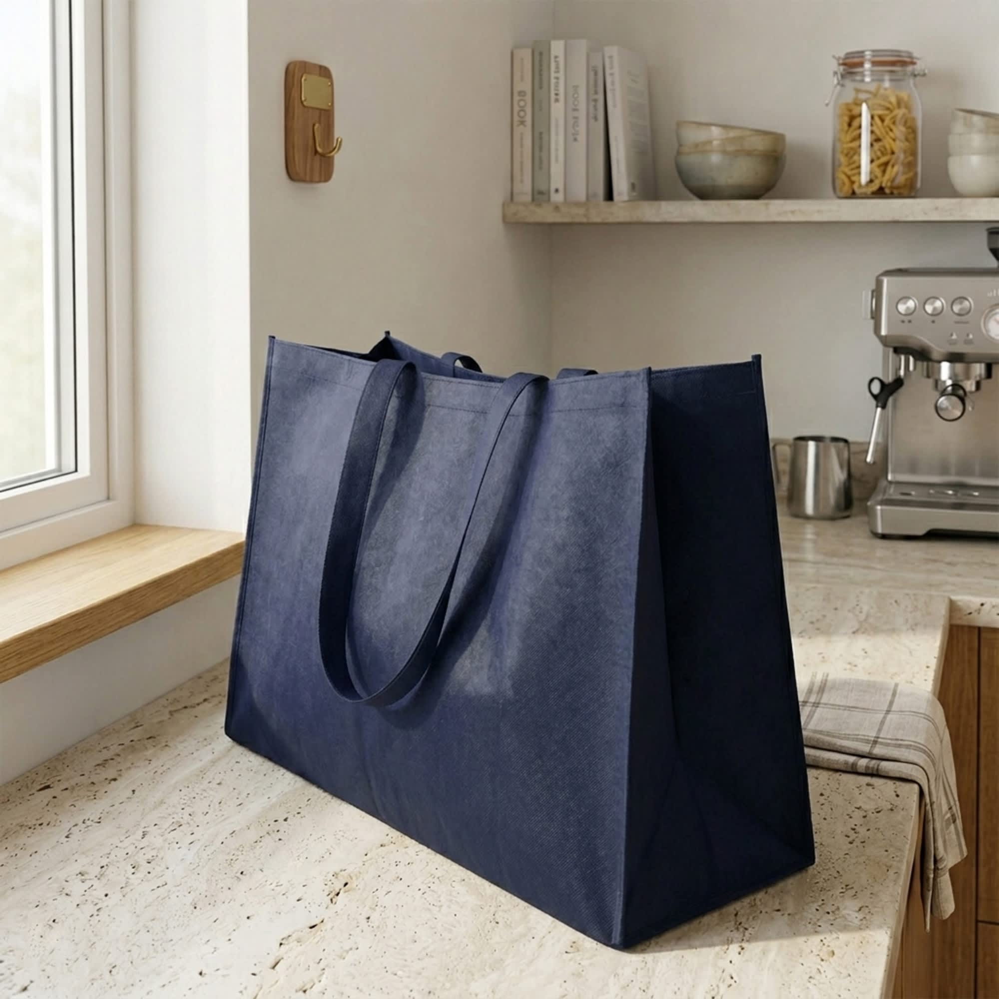 A large navy blue non-woven shopper bag with sturdy handles rests on a kitchen counter. It features a subtle branding logo. Jumbo Non Woven Shopper - Image 1