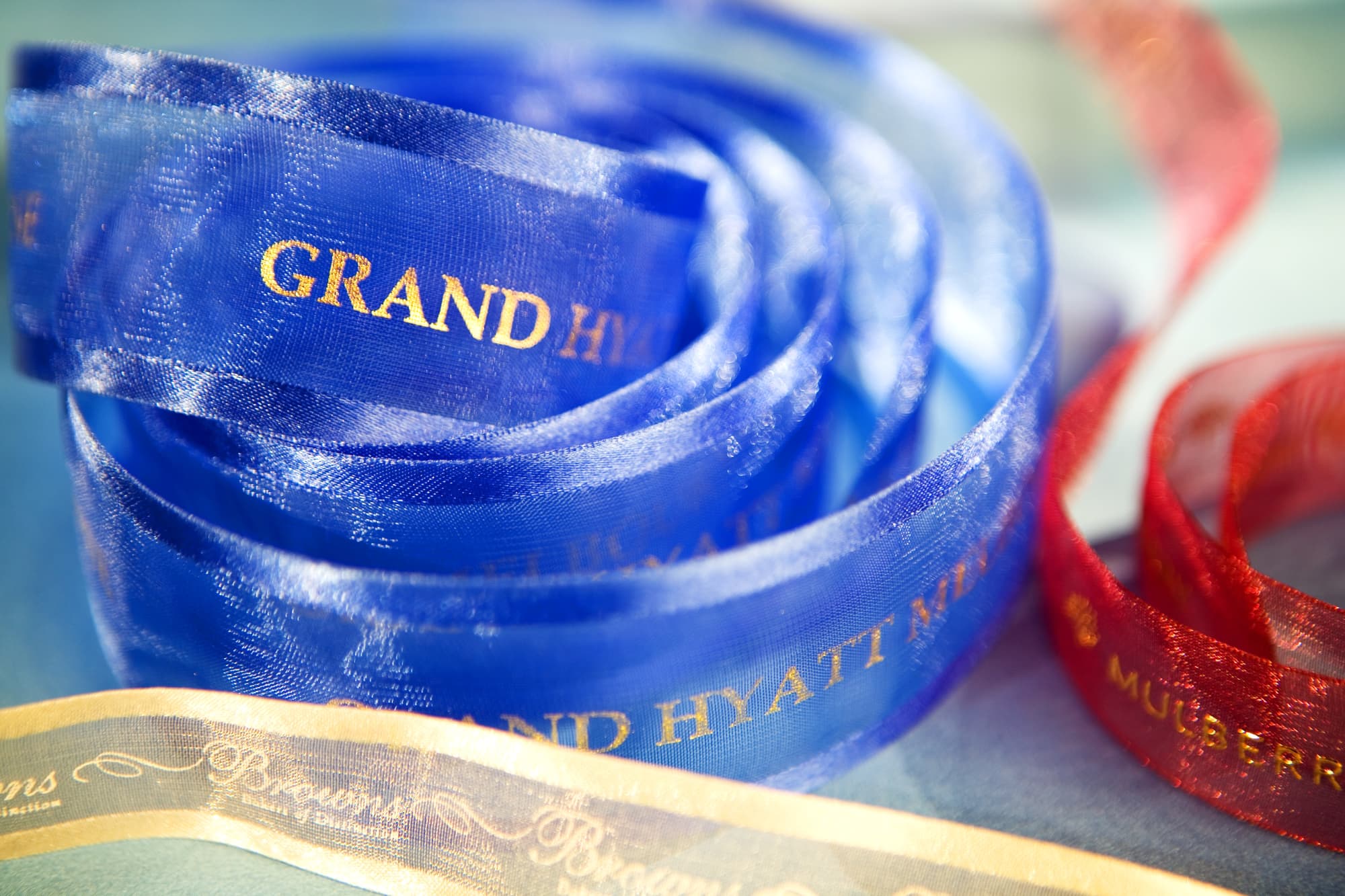 A blue and red organza ribbon with satin edges, featuring a logo and text in gold. Several ribbons are coiled together. Custom Printed Organza Ribbon 38mm Satin Edge - Image 1