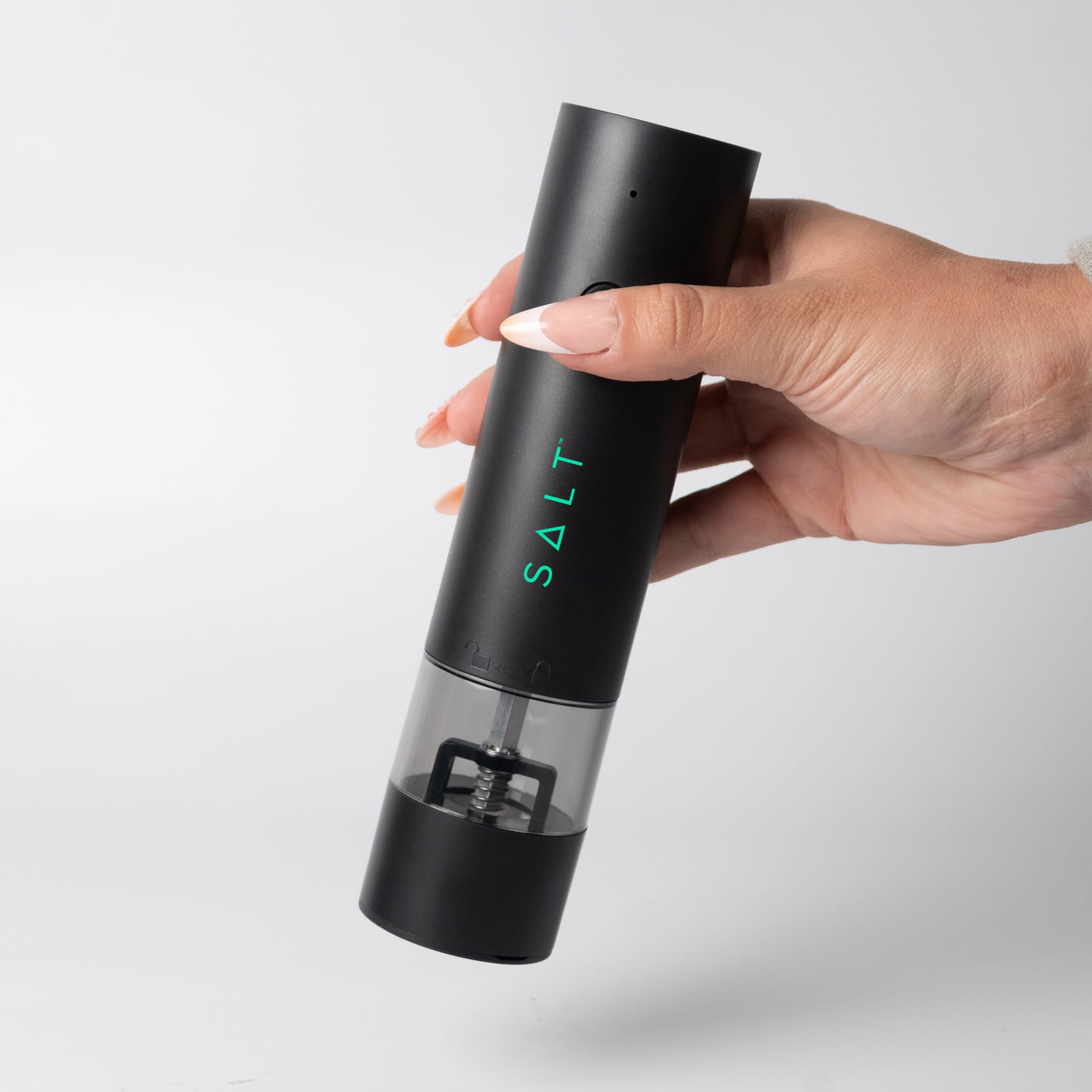 A rechargeable black pepper/salt grinder with a clear base, held in a hand. It features a branding logo. The Seasons Rechargeable Pepper/Salt Grinder - Image 1