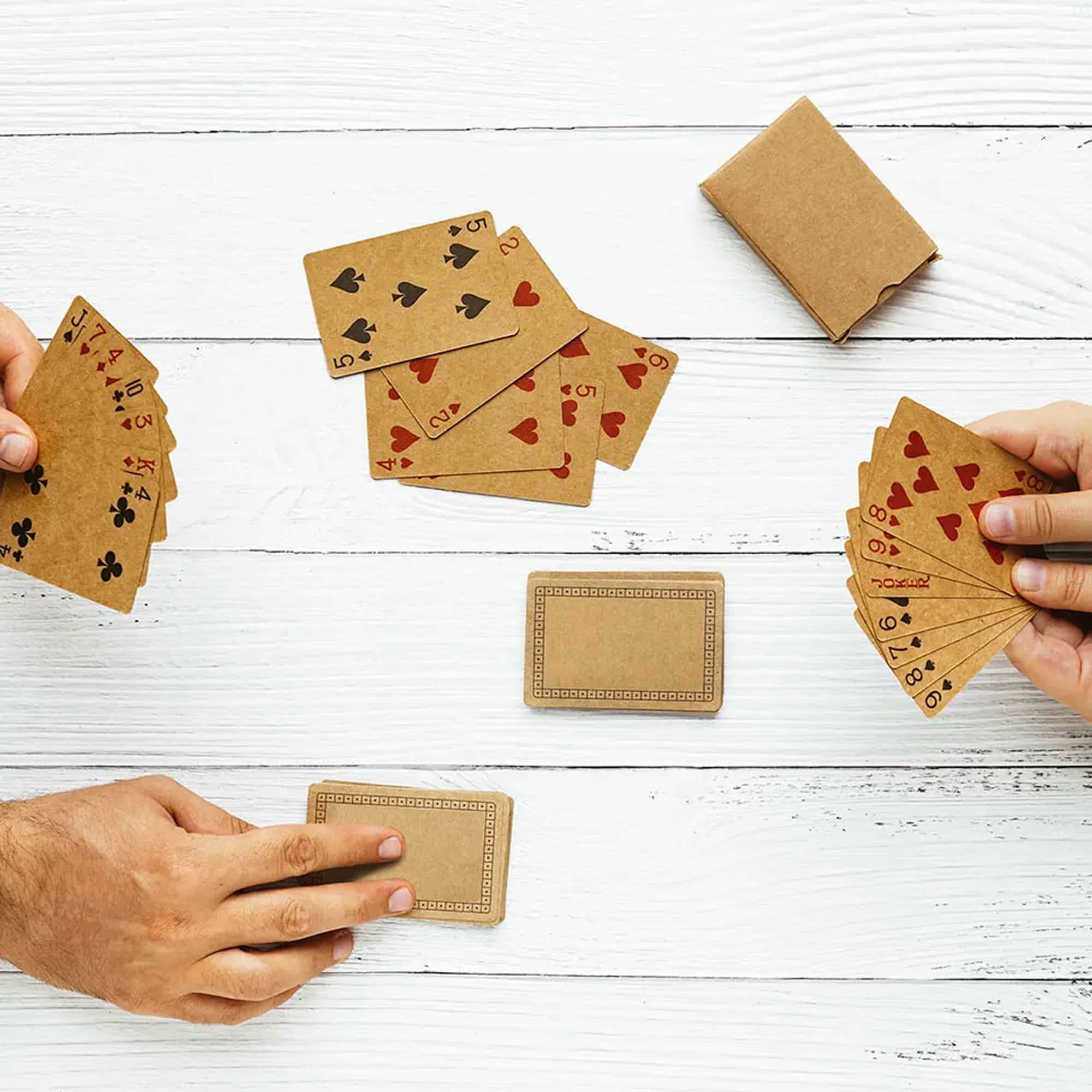 Eco-friendly playing cards in brown recycled material, displayed in hands with a deck and some cards laid out on a wooden surface. Eco Recycled Playing Card - Image 1