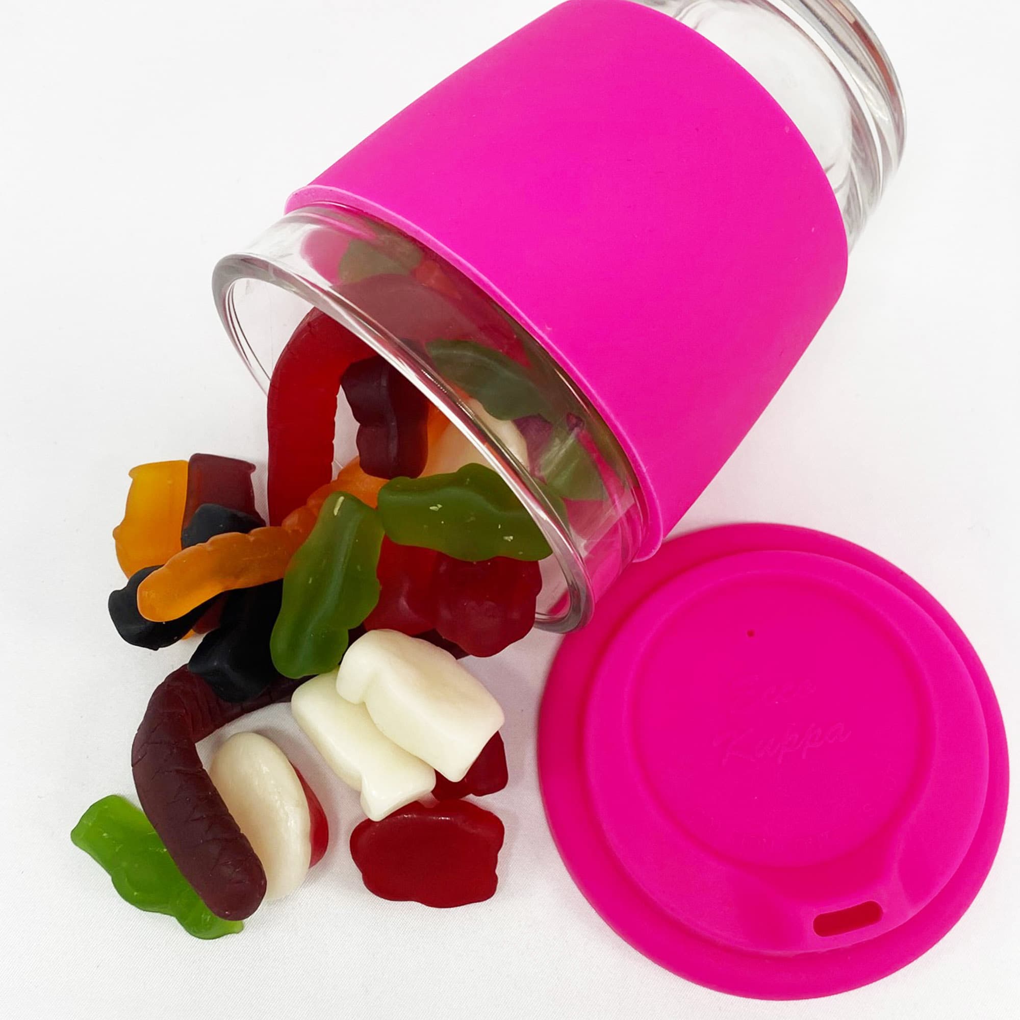 A reusable glass coffee cup with a pink silicone sleeve, filled with assorted jelly lollies spilling out. Jelly Party Mix in Carlo Glass Coffee Cup - Image 1