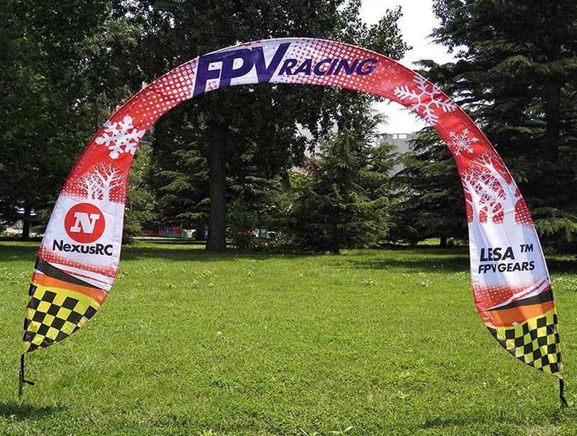 An arch banner in red, white, and black with logos, set in a green grassy area surrounded by trees. Arch Banner 3000mm x 2500mm - Image 1
