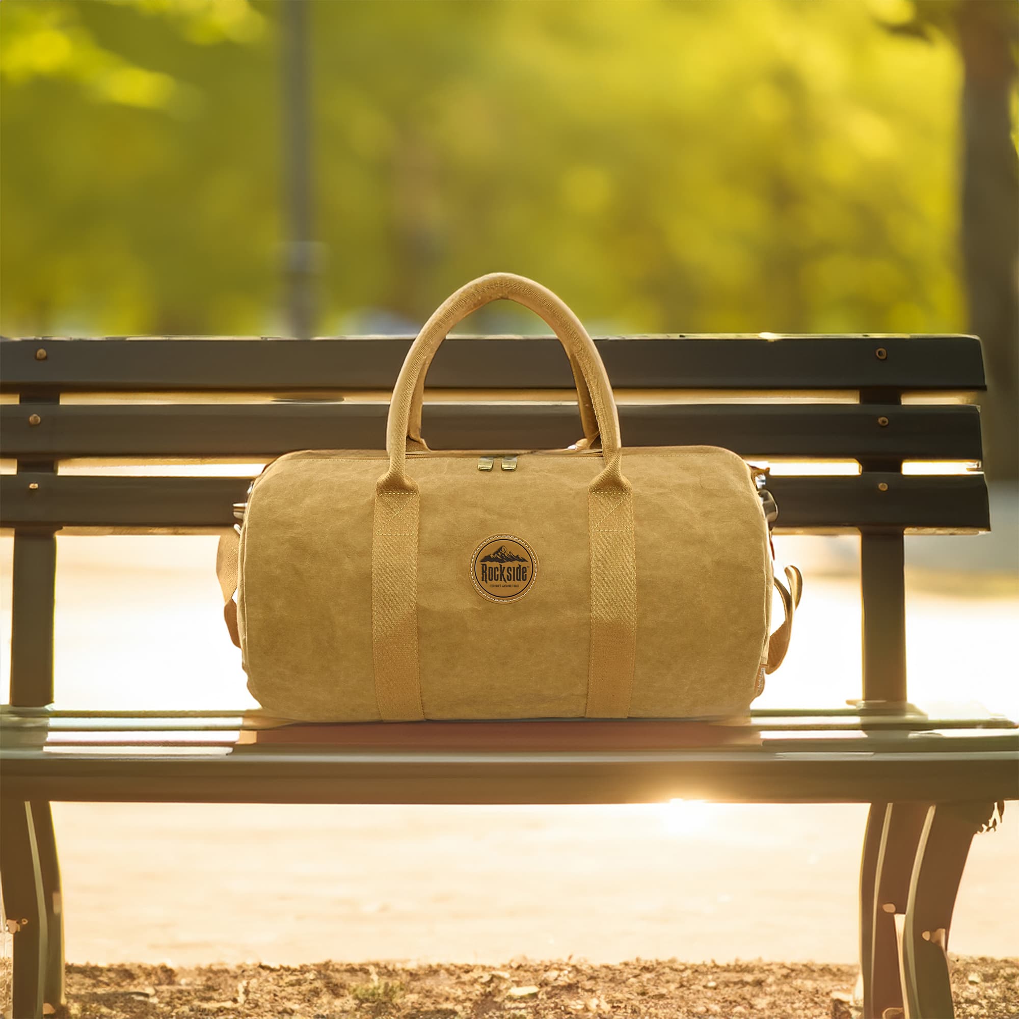 A kraft paper duffle bag in a tan colour sits on a park bench, featuring handles and a logo. Jakob Kraft Paper Duffle Bag - Image 1