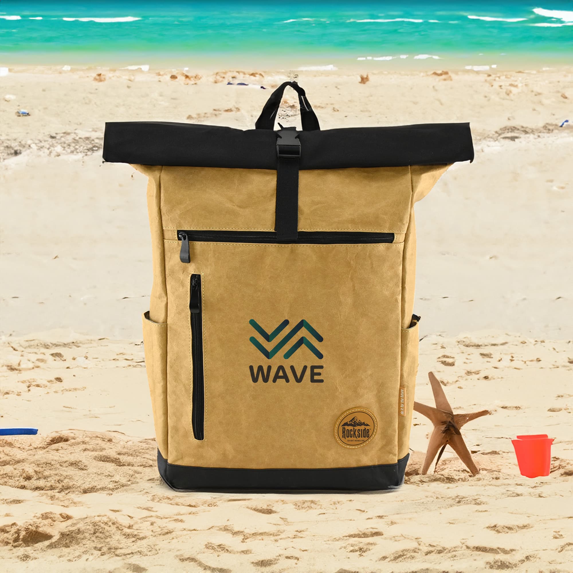 A khaki and black laptop backpack with a logo, placed on a sandy beach setting. Seaside Kraft Paper Laptop Backpack - Image 1