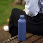A drink bottle in matte navy blue, featuring a sleek design and a logo, resting on a wooden bench.