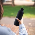 A drink bottle in matte black, featuring a double wall design, held in a person's hand against a blurred outdoor background.
