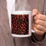 A white mug featuring a design of coffee beans, being held by a person with steam rising from it.