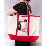 A large canvas tote bag in cream and red, featuring a logo and holding sunglasses and a drink bottle.