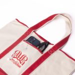 A canvas tote bag in cream and red, featuring a logo and pockets with a phone and sunglasses visible inside.