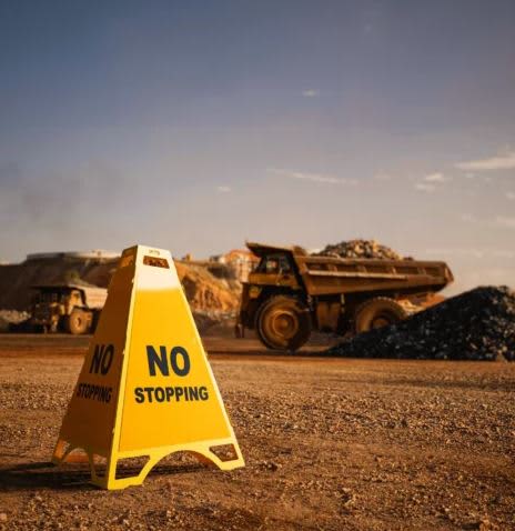 A yellow caution sign displaying "NO STOPPING" on a gravel surface with earthmoving trucks in the background. Metal Pyramid Signage Stand - Image 1