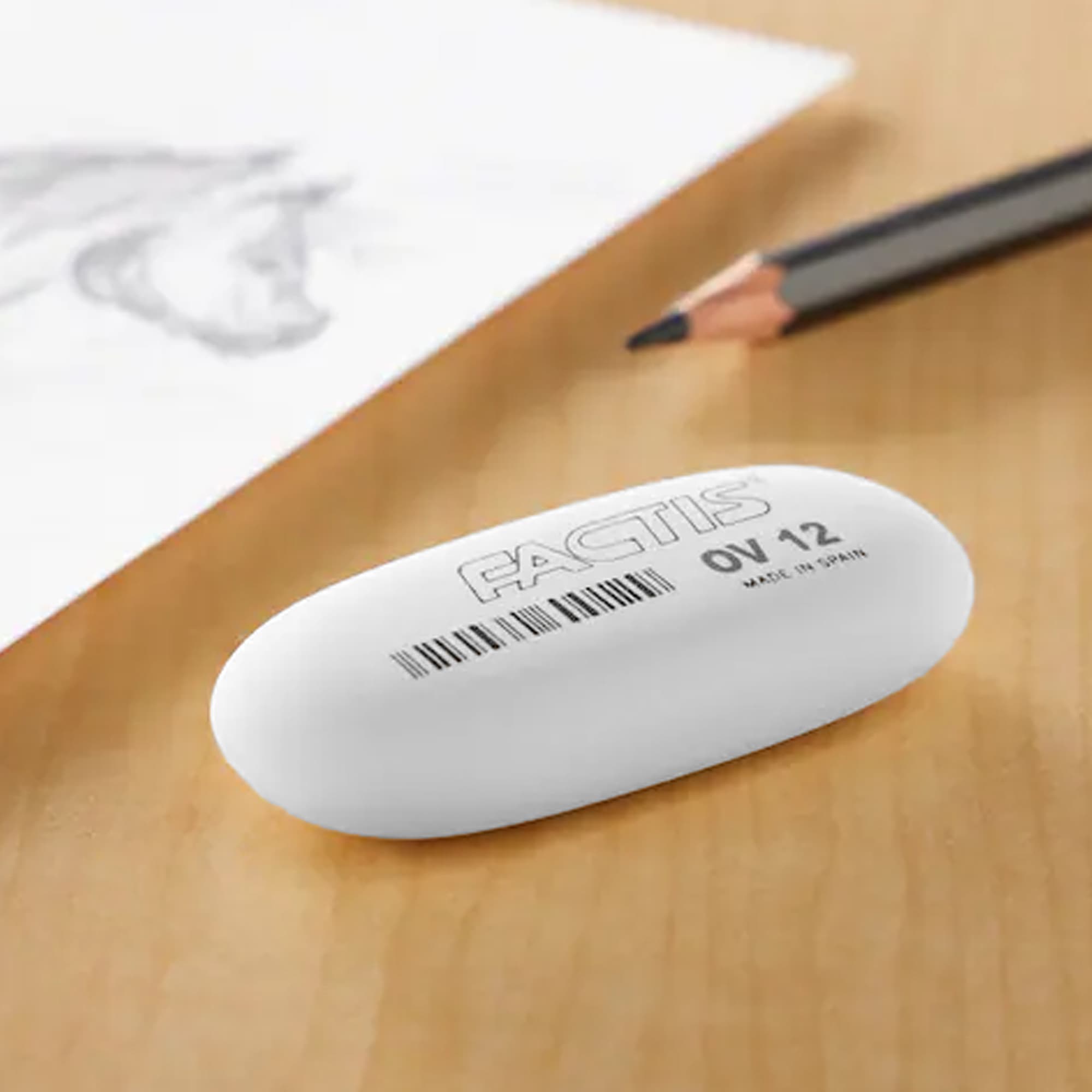A white rubber eraser on a wooden surface beside a drawing and a pencil. The eraser features a logo. Joy Rubber Eraser - Image 1