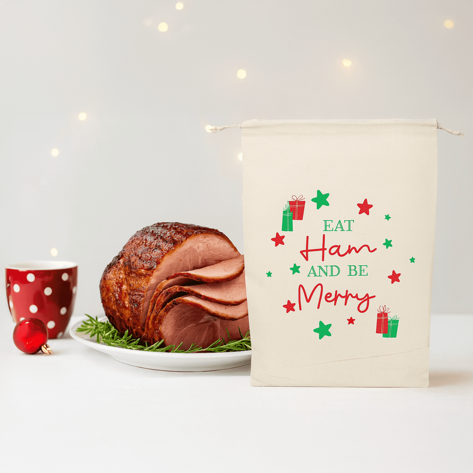 A cream-coloured bag with festive designs next to a glazed ham on a platter and a red polka dot cup. Christmas Ham Bag - Image 1