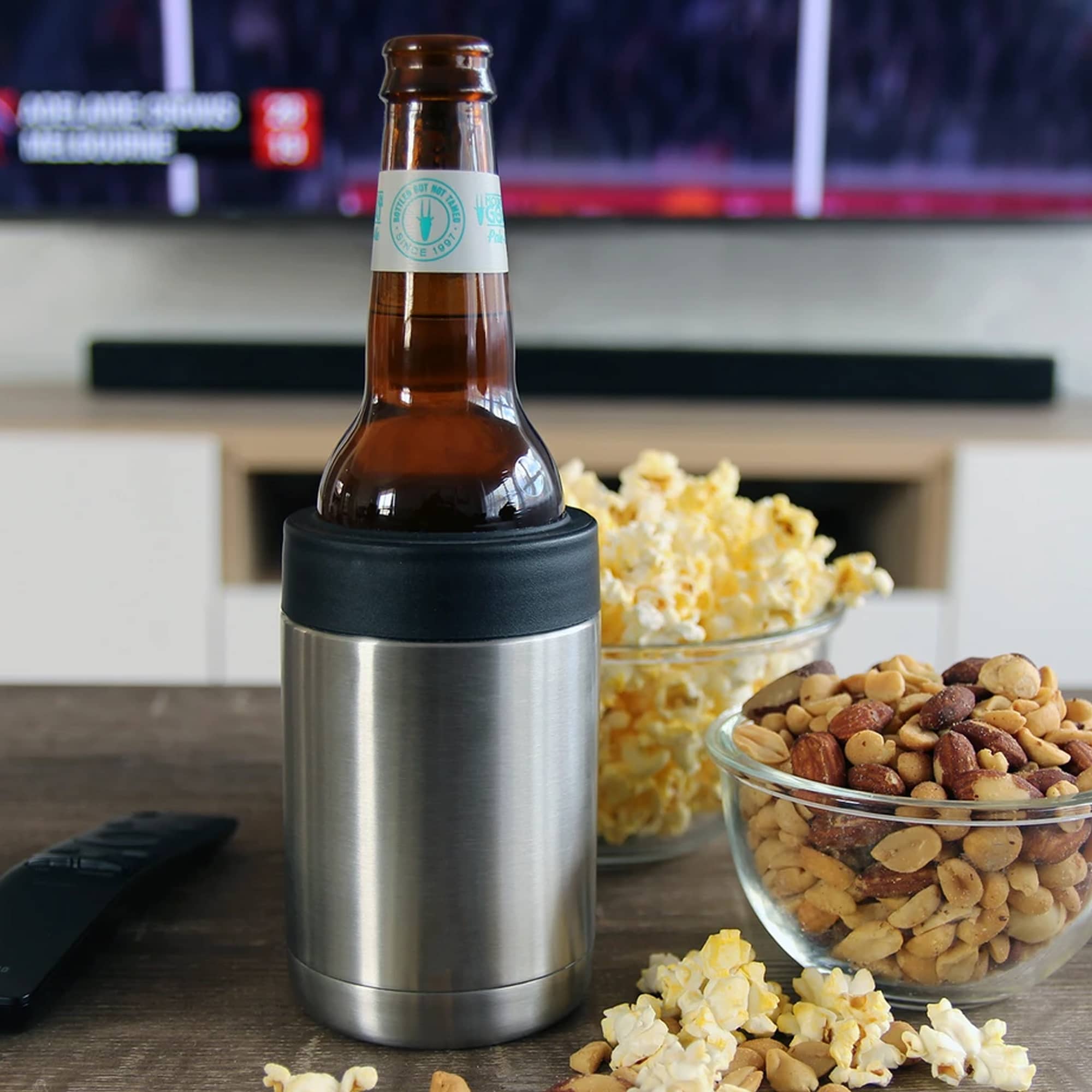 A stainless steel stubby holder in silver with a black silicone base, holding a bottle, surrounded by snacks. Kovina Stainless Steel Stubby Holder - Image 1