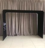 A black fabric arch frame with a tension design, set against a neutral curtain backdrop. Features a logo on one side.