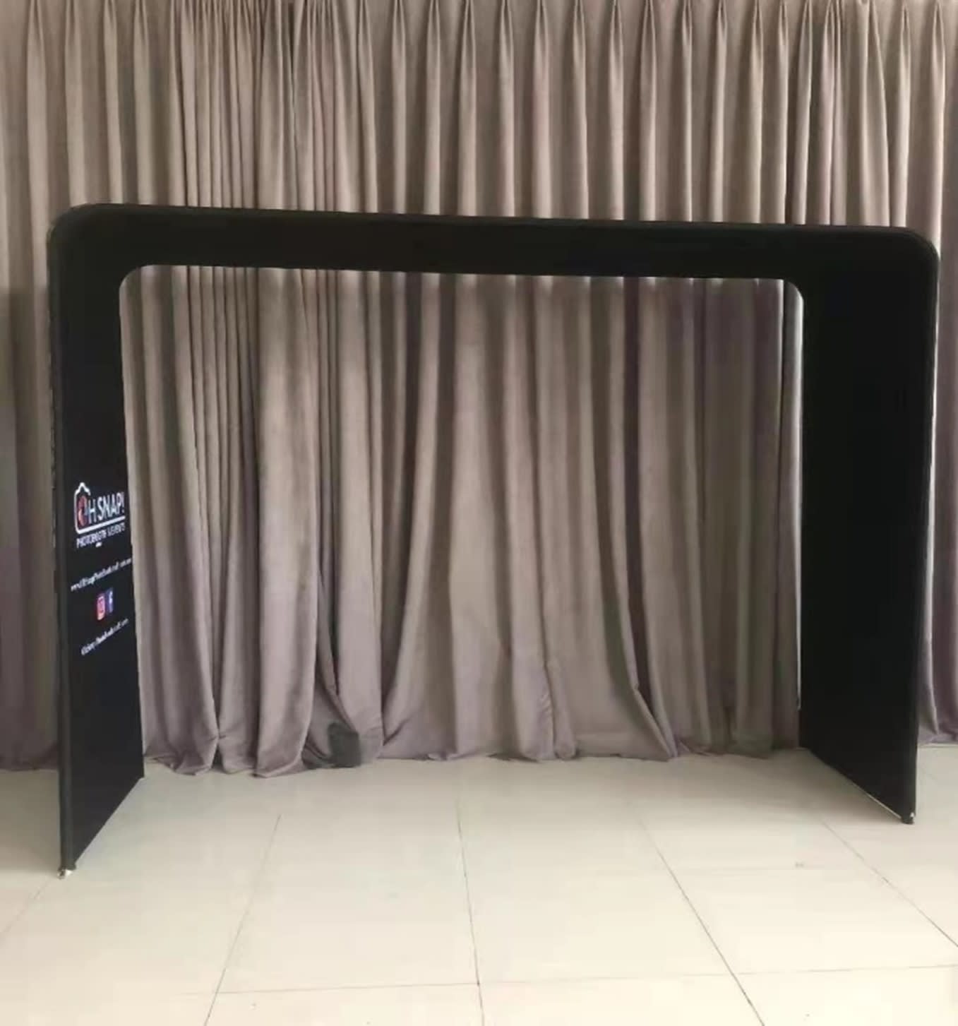 A black fabric arch frame with a tension design, set against a neutral curtain backdrop. Features a logo on one side. A black fabric arch frame with a tension design, set against a neutral curtain backdrop. Features a logo on one side.