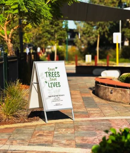 A-frame sign in white and silver displaying a message about saving trees, set in a landscaped outdoor area. Snap Lock A Frame / A-Frame - Image 1