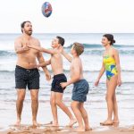 A soft footy ball in vibrant colours is tossed among four people on a beach. The ocean is visible in the background.