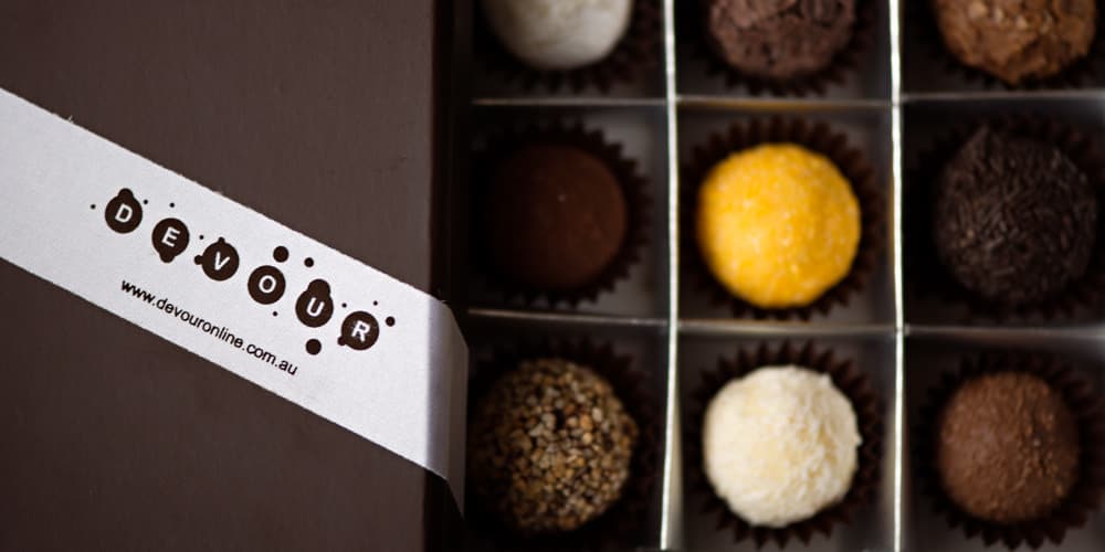 A box of assorted chocolates featuring different colours and textures, with a branded band across the front. Custom Printed Sticky Backed Satin Ribbon 24mm - Image 1