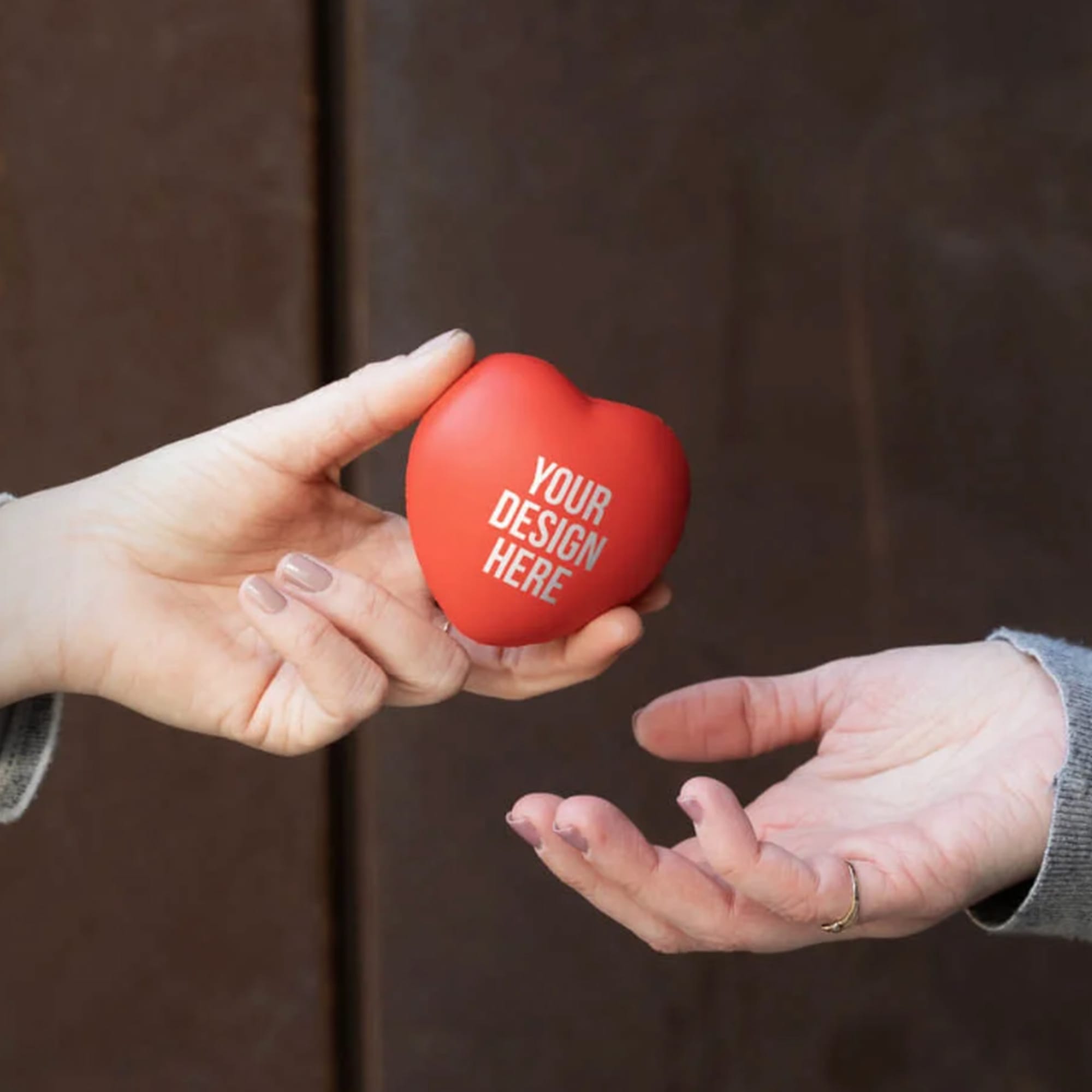 A red heart-shaped stress ball is being handed from one hand to another. The product features a blank area for custom design. Stress Heart - Image 1