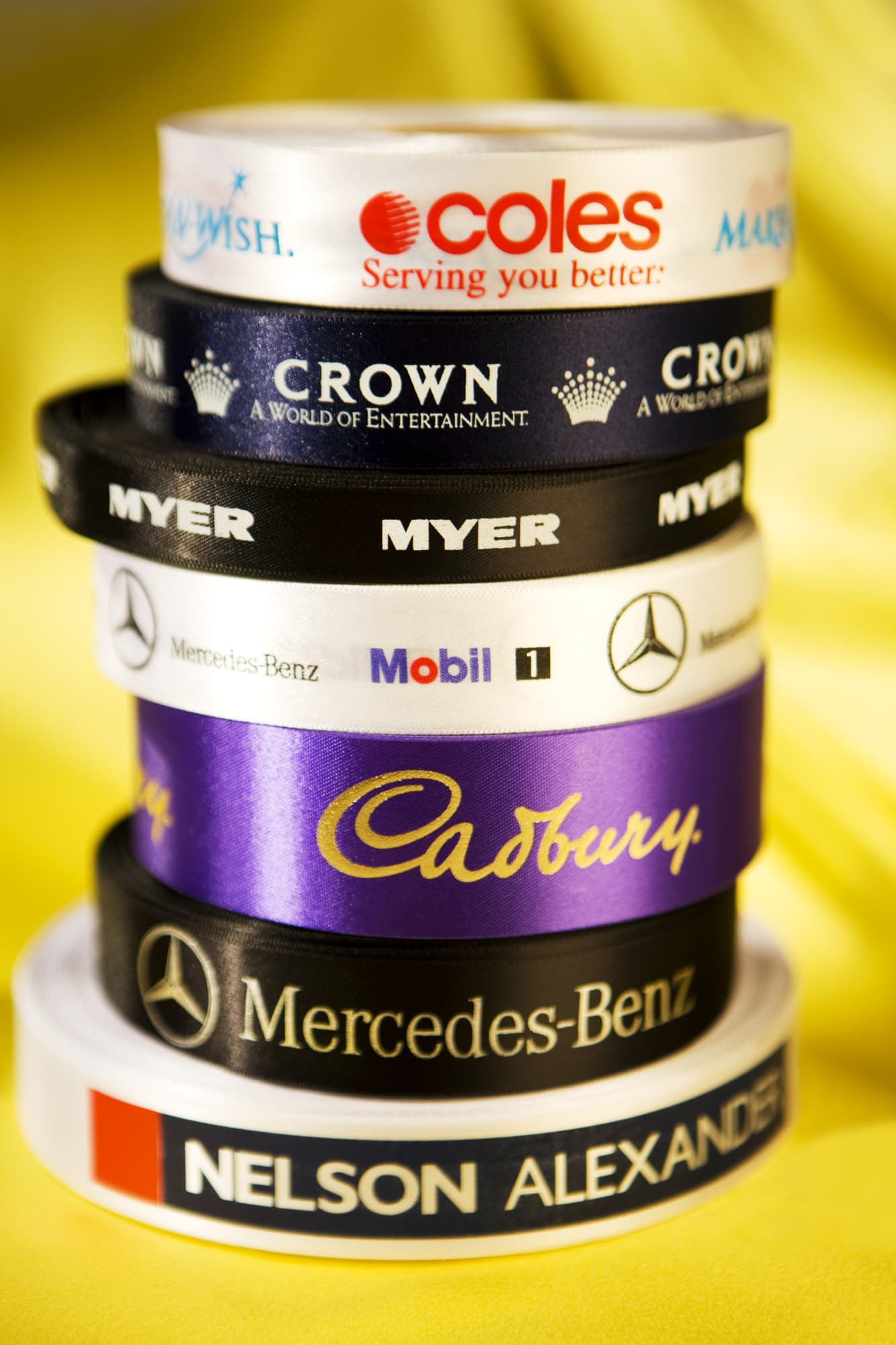 A stack of various satin ribbon rolls in black, white, and purple, featuring logos and branding. Custom Printed Eco Single Sided Satin Ribbon 24mm - Image 1