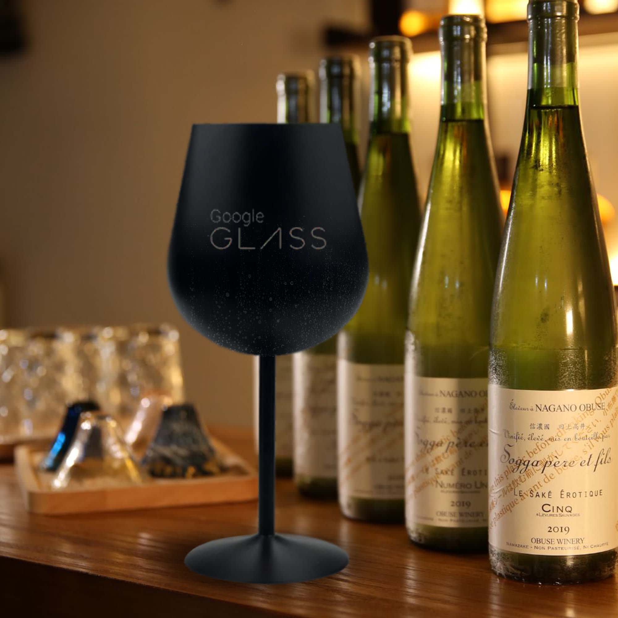A black stainless steel wine glass beside several green wine bottles on a wooden surface. The glass has a branding logo. Bula Vinaka Stainless Steel Wine Glass - Image 1