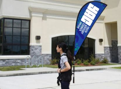 A person carrying a blue teardrop banner backpack with a logo outside a building. Small Double Sided Backpack Tear Drop Banner - Image 1