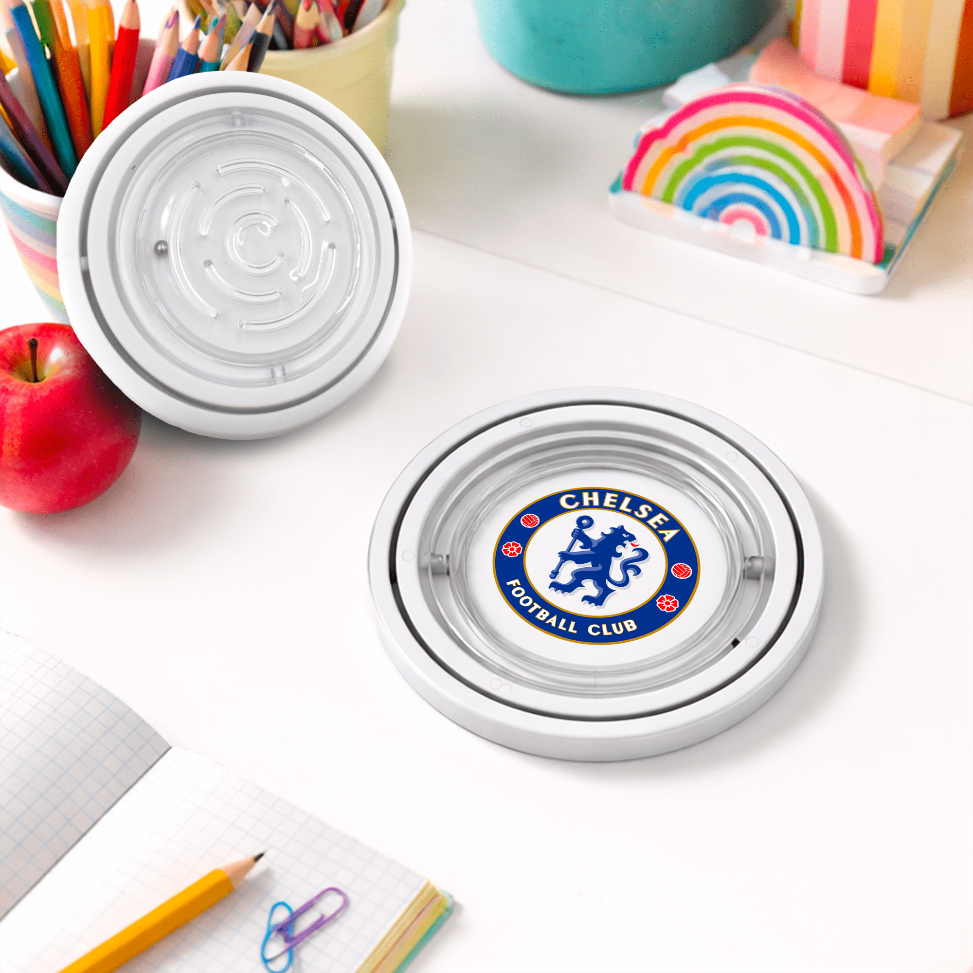 A circular coaster featuring a logo on a table, surrounded by stationery, an apple, and colourful paper. The Amazing Fidget - Image 1