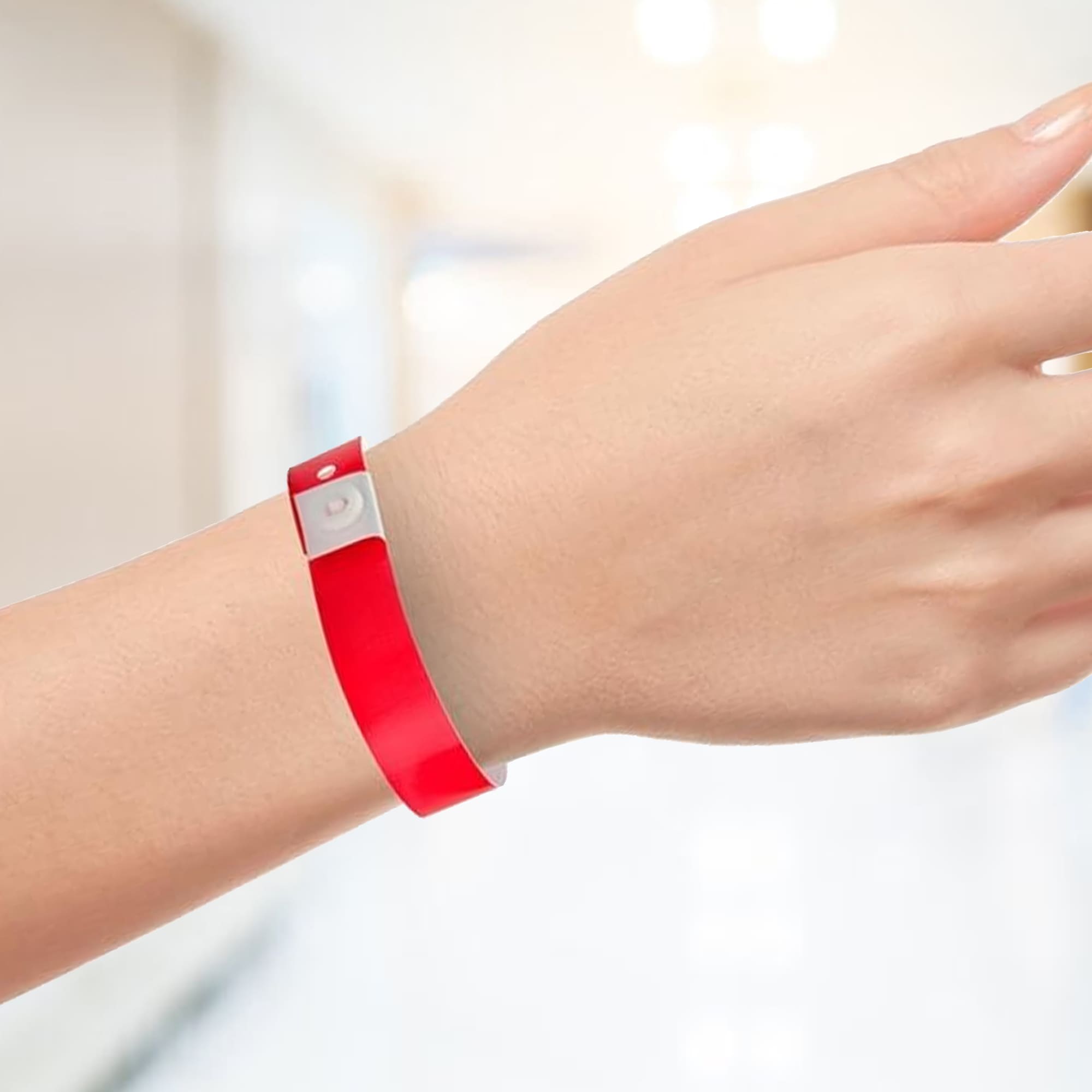 A red plastic wristband is displayed on a hand, featuring a silver fastening. The background is blurred and light-filled. Code Plastic Wrist Band 16mm - Image 1