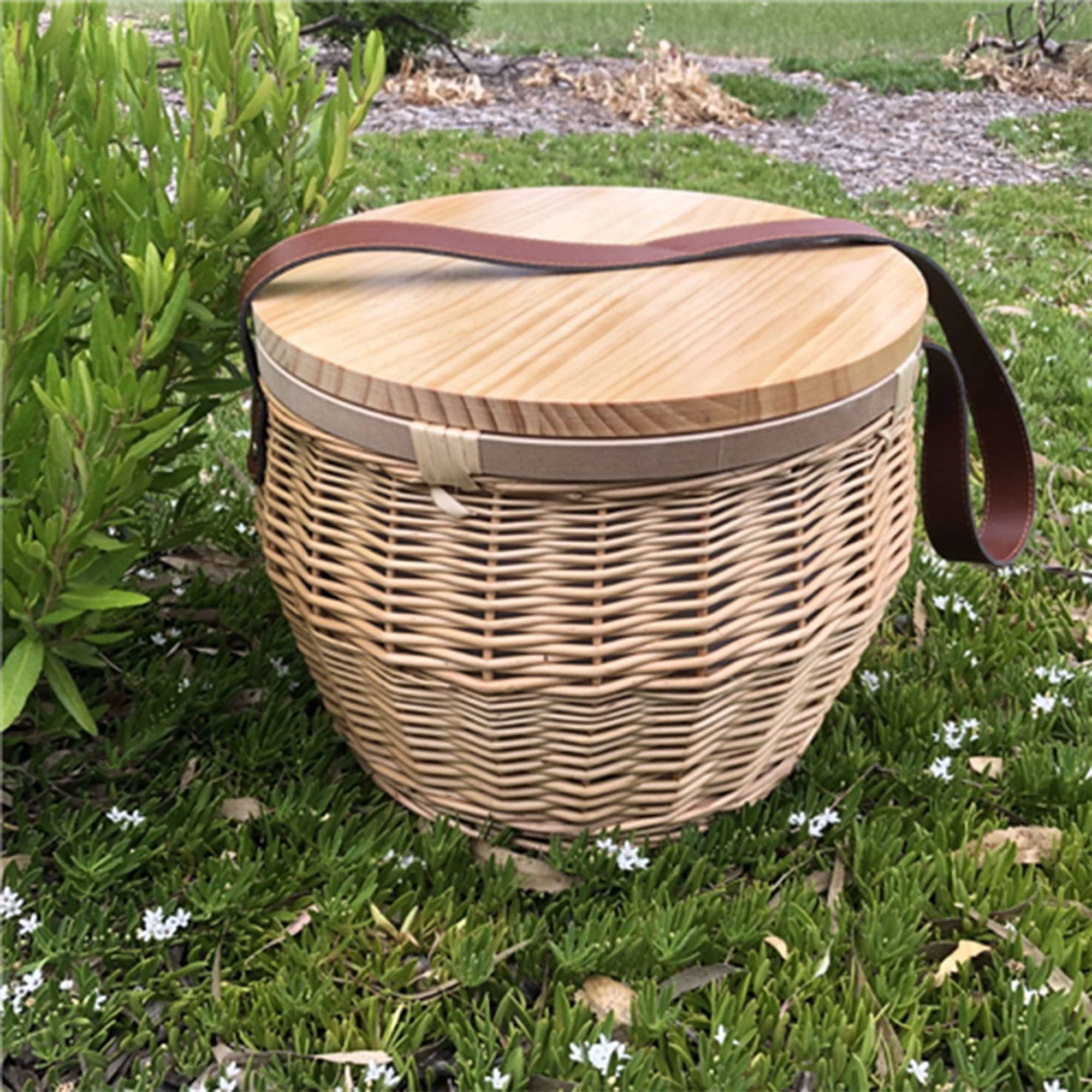 A round wicker picnic cooler basket with a wooden lid and a leather strap, set on grass with white flowers. Scotch Wicker Picnic Cooler Basket Round - Image 1