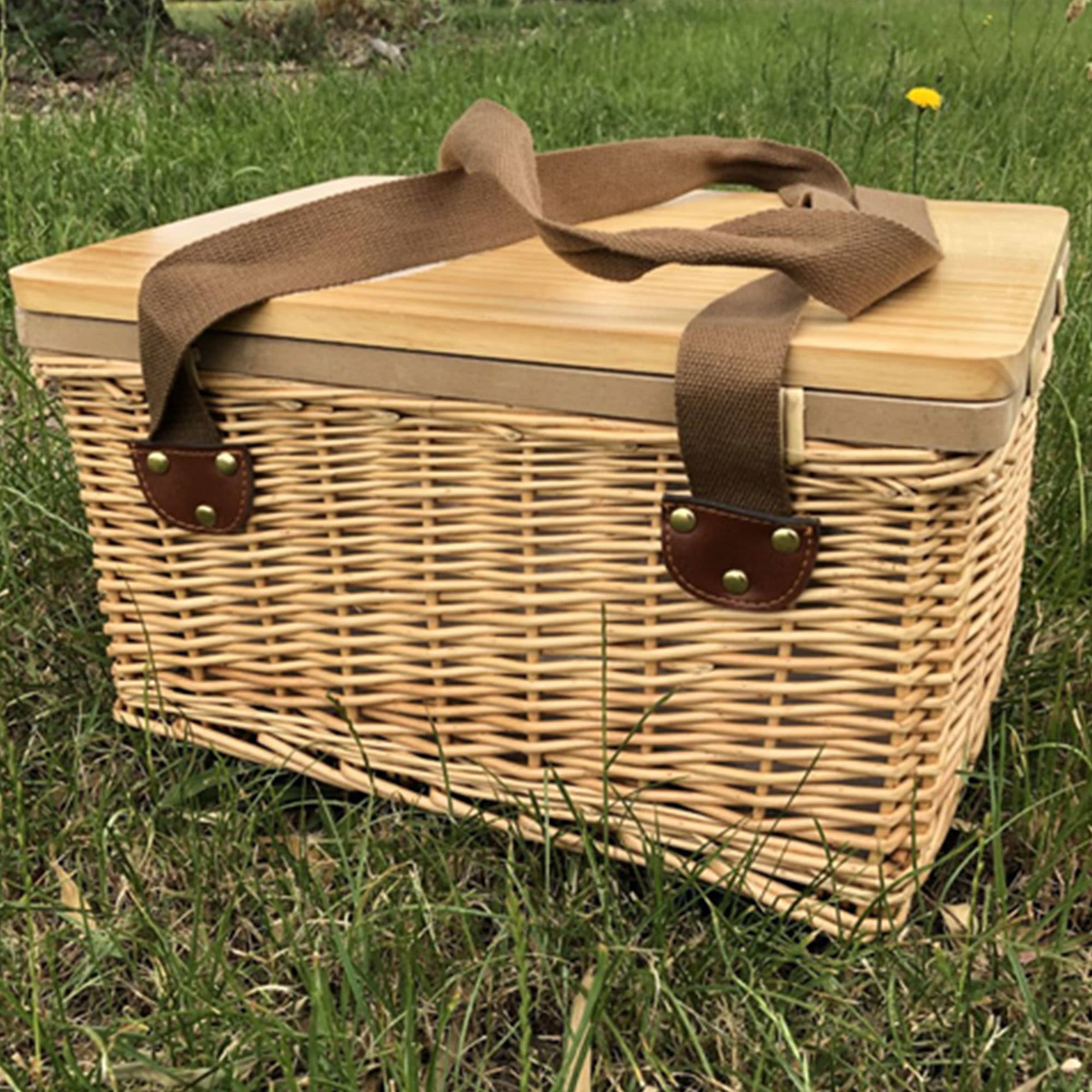 A wicker picnic cooler basket in natural tones with a bamboo lid and brown fabric handles, resting on grass. Gold Coast Wicker Picnic Cooler Basket Square - Image 1