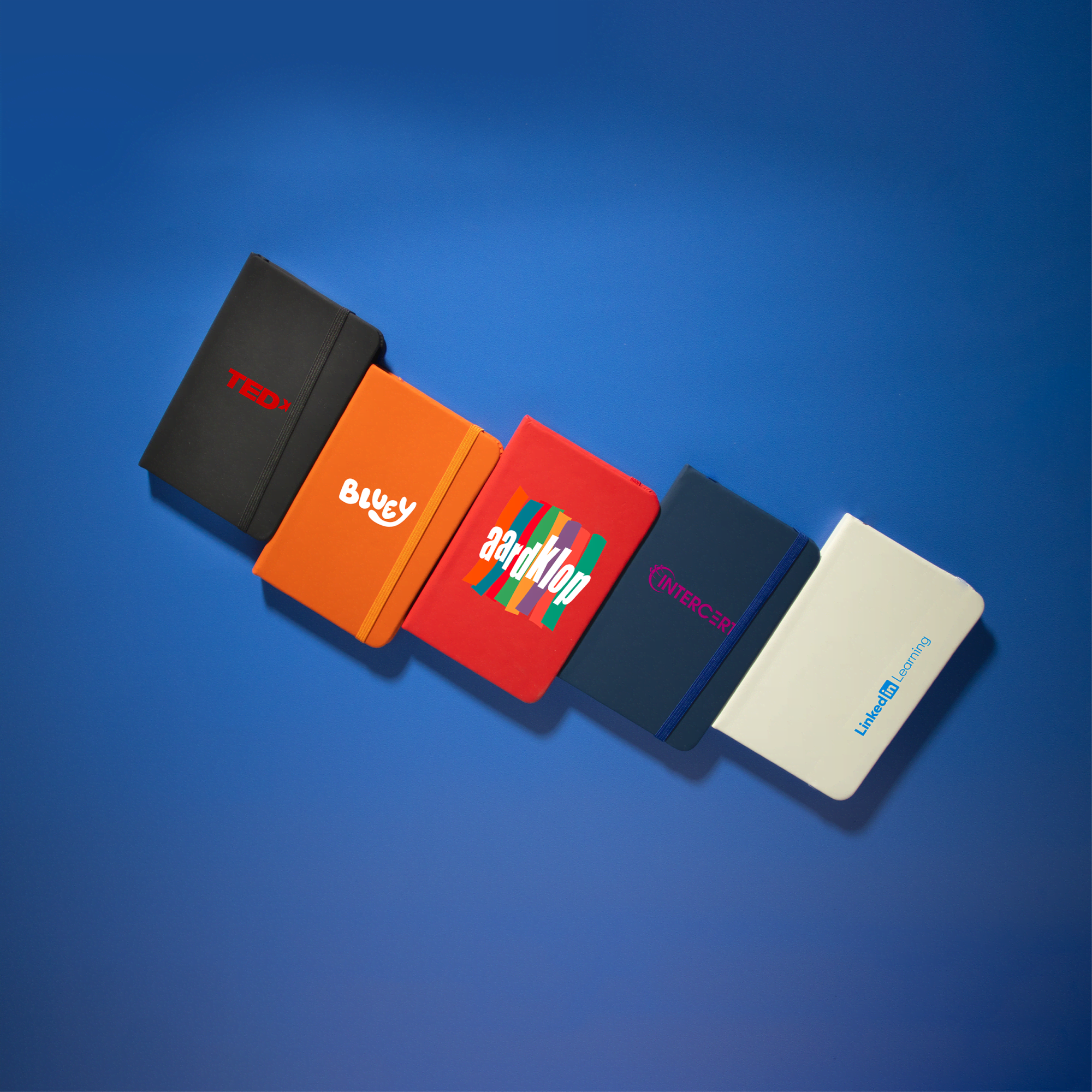 A collection of five Minichroma A6 Notebooks in various colours: red, orange, blue, navy, and white, each featuring a logo. Minichroma A6 Notebook - Image 1