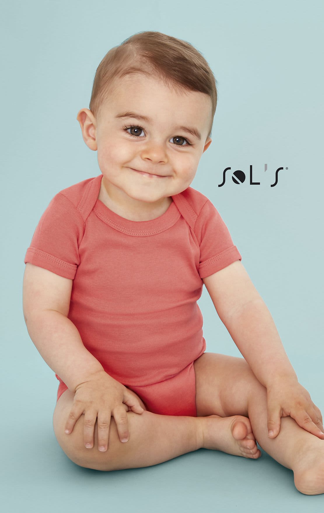 A baby bodysuit in coral pink, featuring short sleeves, displayed by a smiling child against a light blue background. BABY BODYSUIT Bambino - Image 1