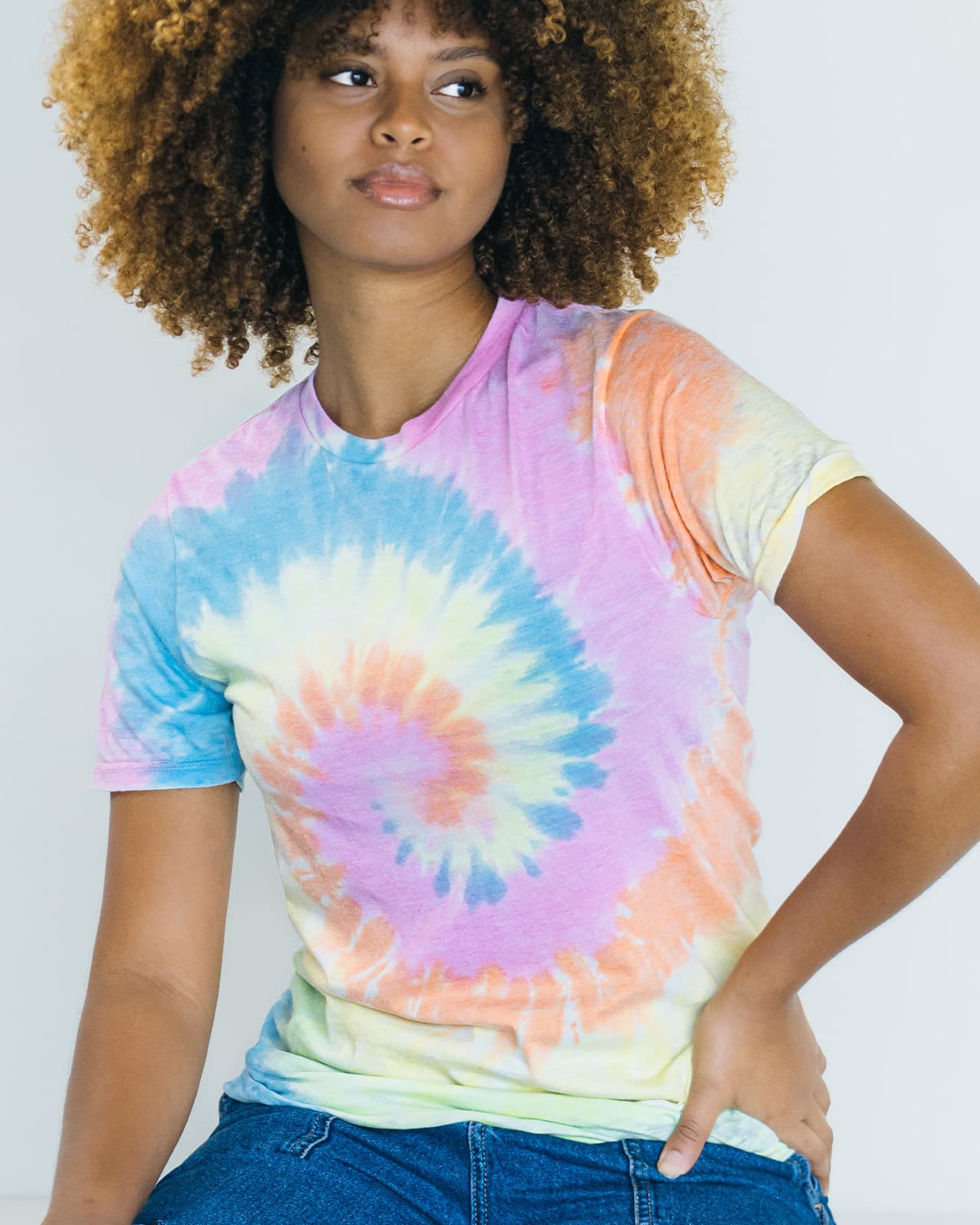 A woman models a Colortone® tie dye adult T-shirt featuring a spiral pattern in pastel colours of blue, pink, yellow, and green. Colortone® Tie Dye Adult T-Shirt - Image 1