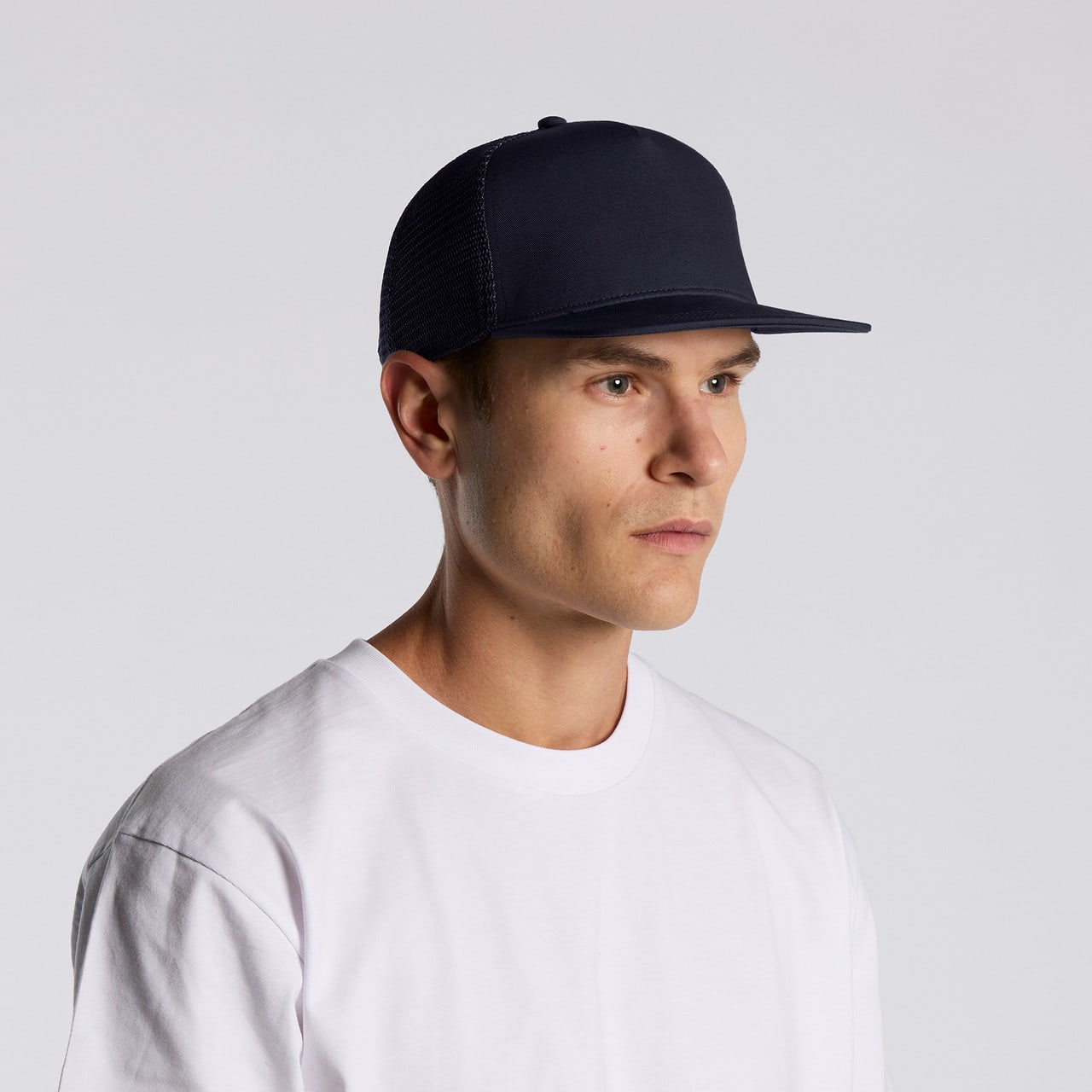 A navy trucker cap with a flat brim, featuring a logo, worn by a model in a white T-shirt. Stock Trucker Cap - Image 1
