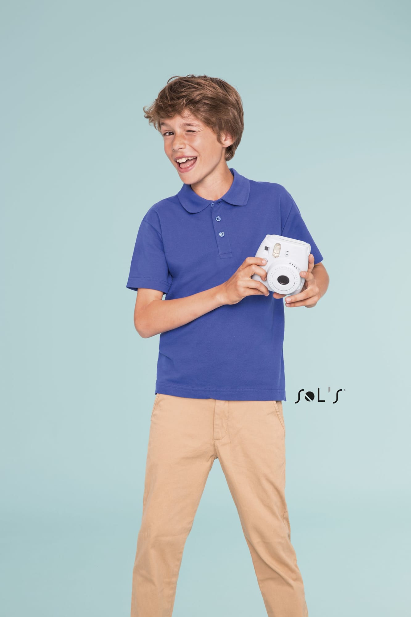 A boy in a blue polo shirt smiles while holding a camera, standing against a light background. Polo shirt KIDS 100% combed ring spun cotton SUMMER II - Image 1