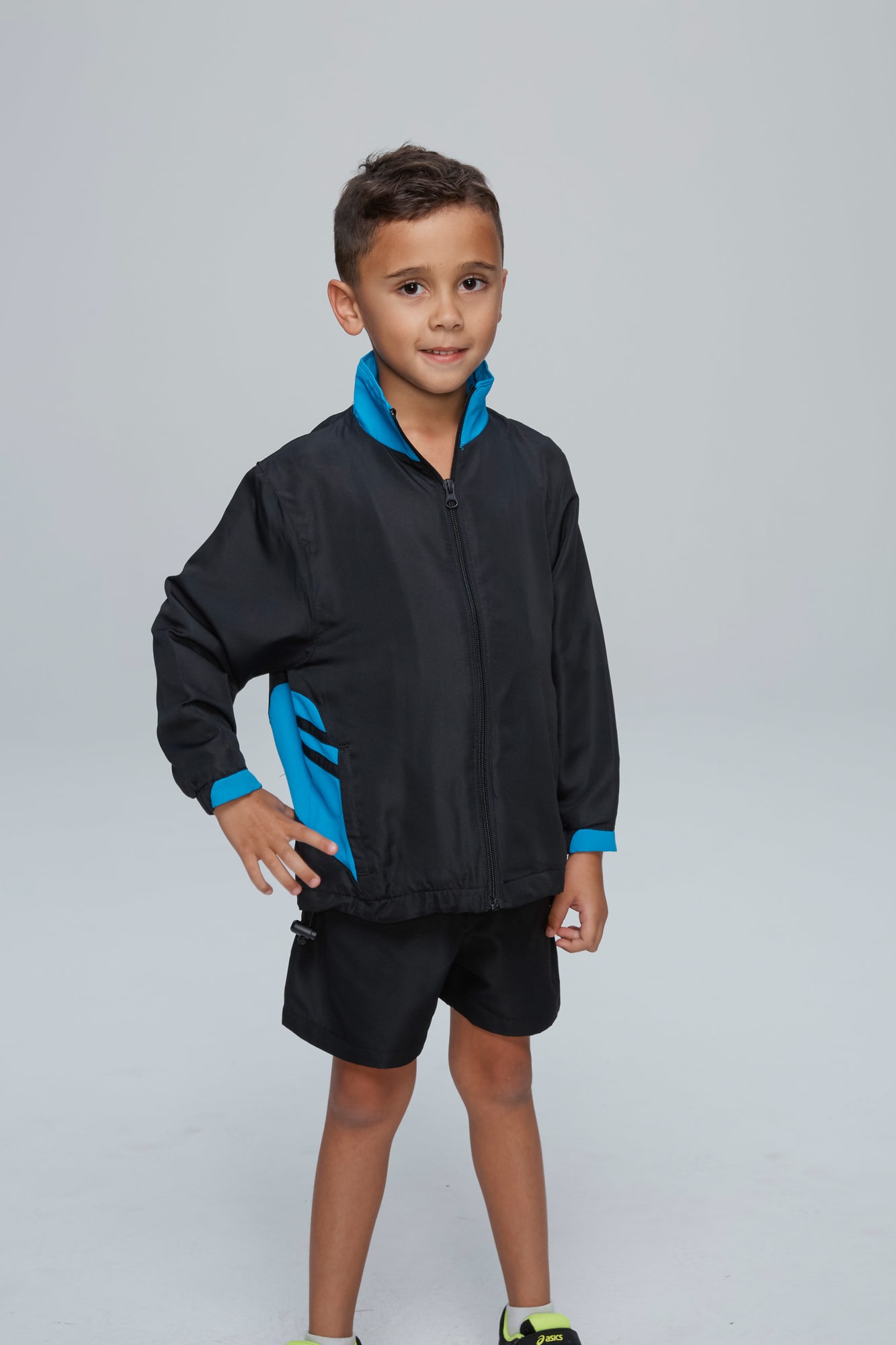 A child wearing a black tracktop with blue accents, paired with black shorts, stands confidently. TASMAN KIDS TRACKTOPS RUNOUT - Image 1