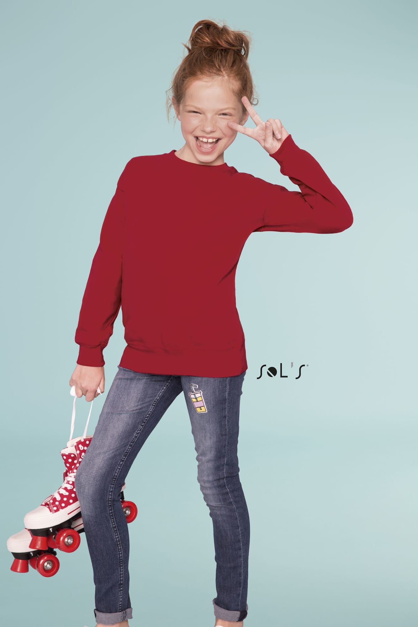A girl in a red sweatshirt poses with roller skates while standing in a light blue background. The outfit features a logo. Windcheater / Sweat shirt KIDS SUPREME - Image 1