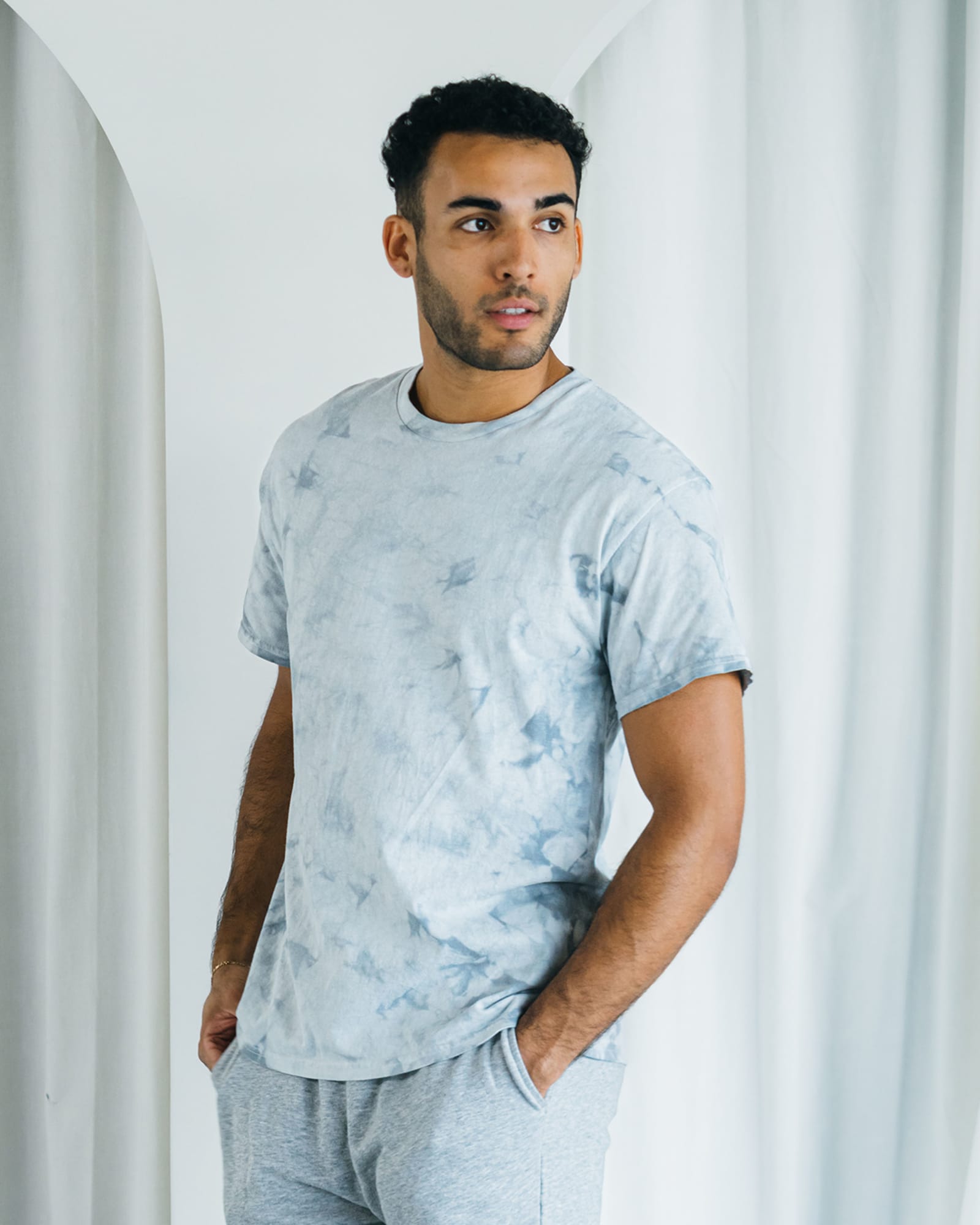 A man wearing a grey tie-dye t-shirt and light grey shorts, standing against a soft, neutral background. Colortone® Crystal Wash Adult T-Shirt - Image 1