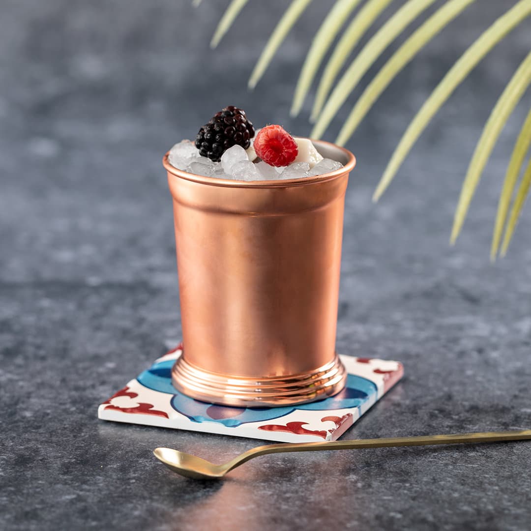 A copper drink cup filled with ice and garnished with blackberries and raspberries, set on a patterned coaster. Riberton - Image 1