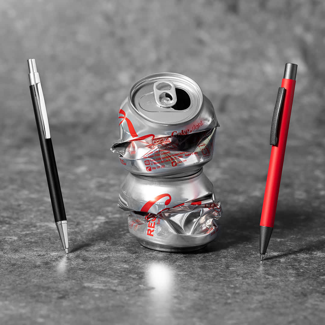 A crushed silver soft drink can sits between a black pen and a red pen on a textured background. The image is monochrome with a pop of colour. Brincio - Image 1