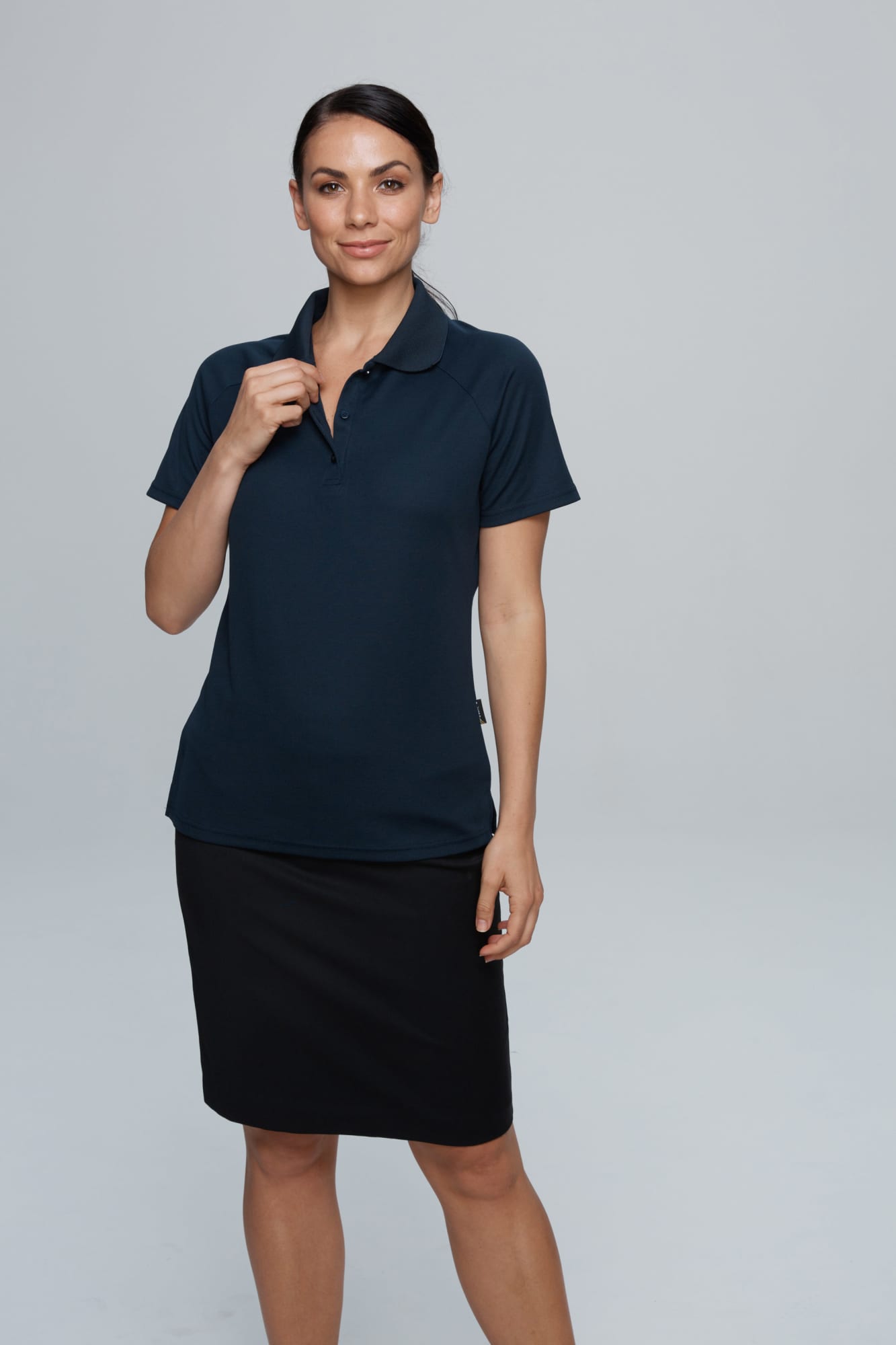 A woman poses in a navy blue polo shirt paired with a black skirt. The shirt features a logo. KEIRA LADY POLOS - Image 1