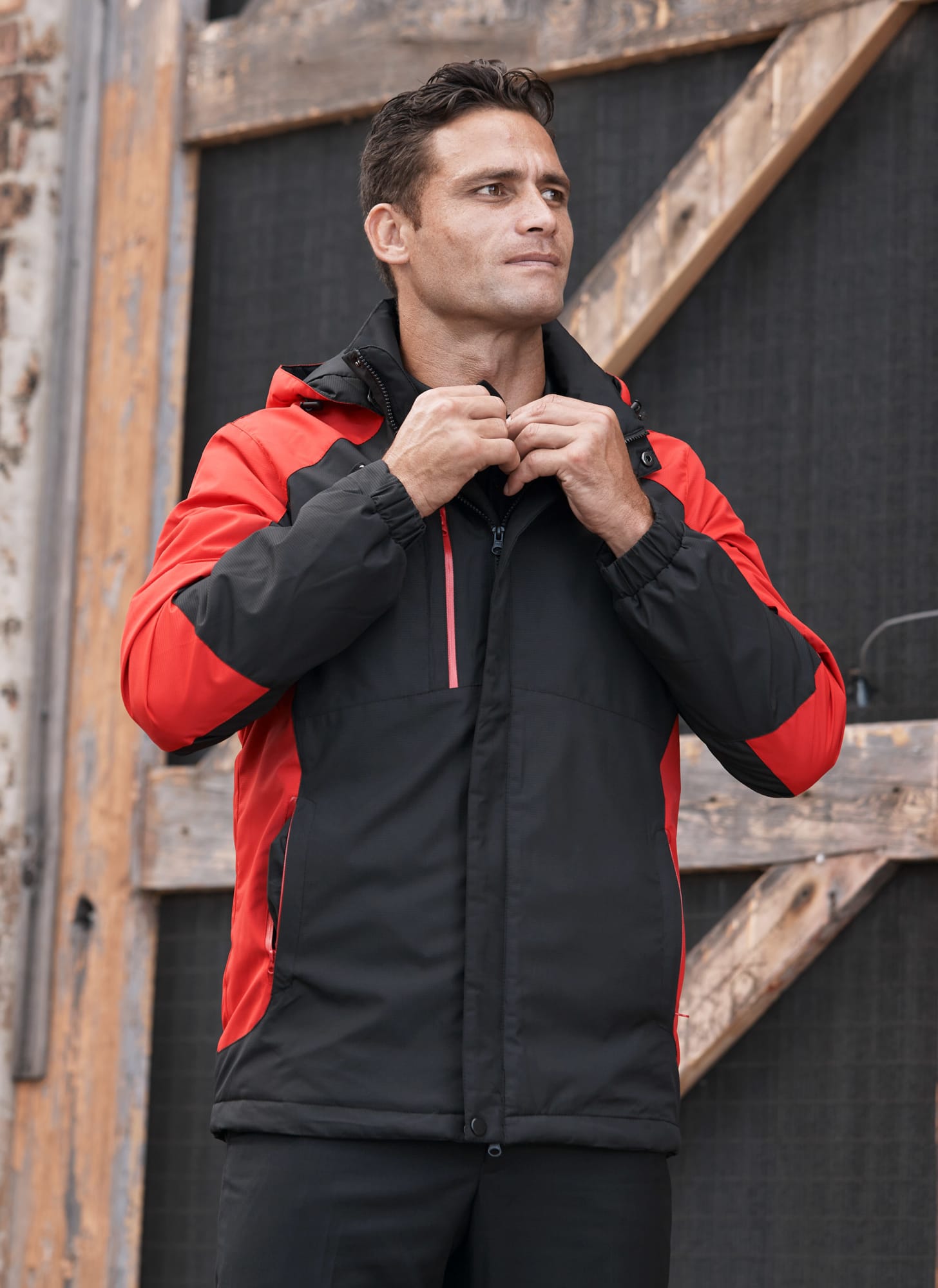 A man wearing a black and red jacket, standing against a rustic wooden backdrop. The jacket has a hood and zippers. NAPIER MENS JACKETS - Image 1