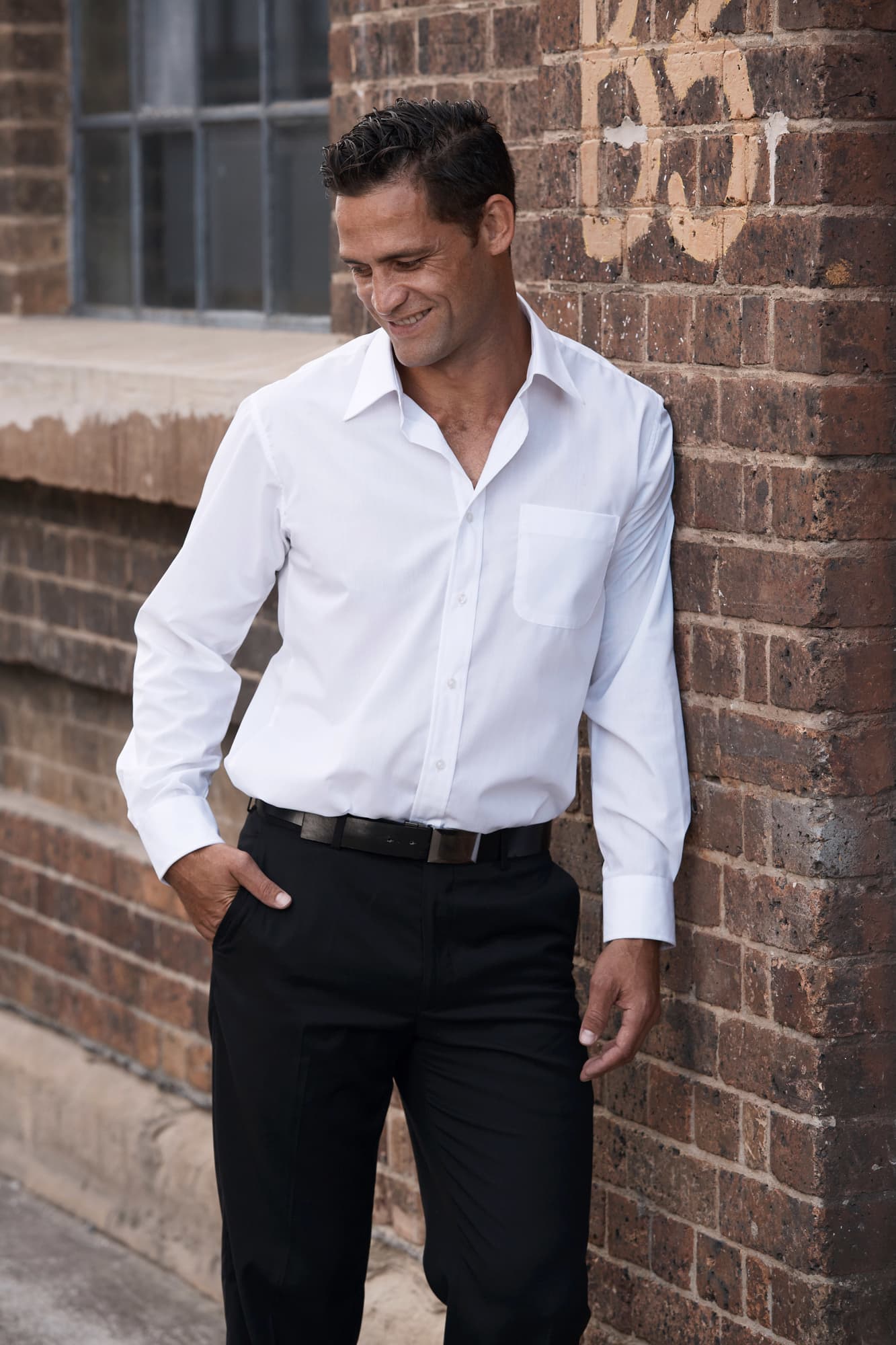 A long sleeve white button-up shirt on a man, paired with black trousers, against a brick wall background. KINGSWOOD MENS SHIRT LONG SLEEVE - Image 1