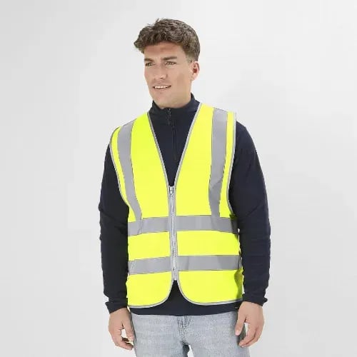 A high-visibility safety vest in bright yellow with reflective grey stripes, worn by a man standing against a plain backdrop. Jestol - Image 1